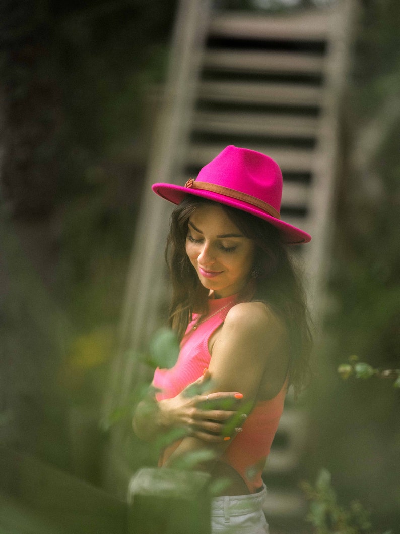 Puede incluir: Una mujer con un sombrero rosa y un top de tirantes coral. Est&aacute; de pie frente a una escalera de madera y vegetaci&oacute;n verde.