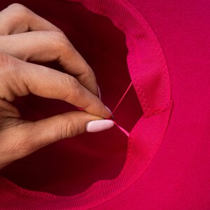 Puede incluir: Primer plano de un sombrero de fieltro rosa con una cinta rosa cosida alrededor del interior del ala. Una mano tira de la cinta para ajustar el tama&ntilde;o del sombrero.