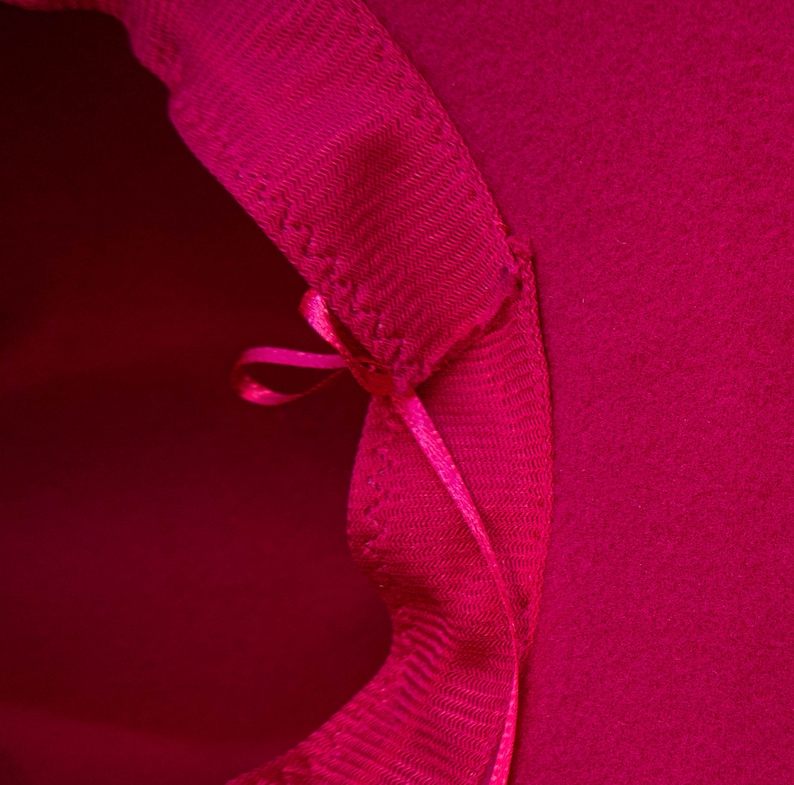 Puede incluir: Primer plano del interior de un sombrero rosa brillante. El sombrero tiene una cinta cosida en la costura interior, atada en un lazo.