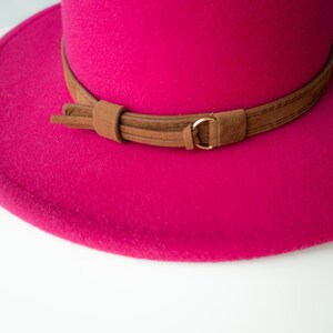 Puede incluir: Un sombrero fedora de fieltro rosa brillante con una banda de cuero marr&oacute;n y una hebilla dorada.