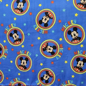 K&ouml;nnte beinhalten: Blaue Fleece-Decke mit einem sich wiederholenden Muster von Mickey Mouse in einem gelben Kreis mit dem Wort "MICKEY" um den Kreis herum gedruckt. Der Hintergrund ist blau mit gelben Sternen.