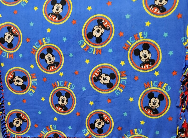 K&ouml;nnte beinhalten: Blaue Fleece-Decke mit einem sich wiederholenden Muster von Mickey Mouse in einem gelben Kreis mit dem Wort "MICKEY" um den Kreis herum gedruckt. Der Hintergrund ist blau mit gelben Sternen.