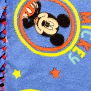 K&ouml;nnte beinhalten: Blaue Fleece-Decke mit einem sich wiederholenden Muster von Mickey Mouse in einem gelben Kreis mit roten und orangen Akzenten. Die Decke ist mit gelben und roten Sternen &uuml;bers&auml;t.
