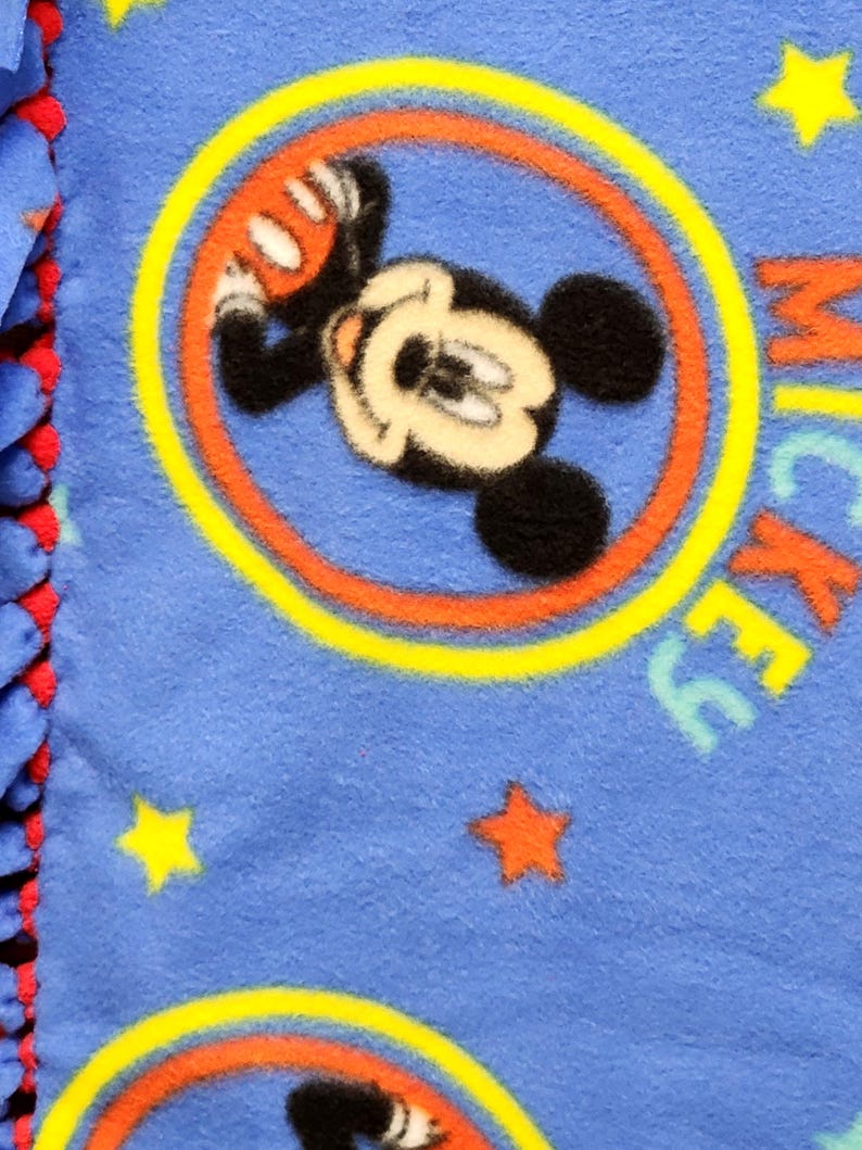 K&ouml;nnte beinhalten: Blaue Fleece-Decke mit einem sich wiederholenden Muster von Mickey Mouse in einem gelben Kreis mit roten und orangen Akzenten. Die Decke ist mit gelben und roten Sternen &uuml;bers&auml;t.
