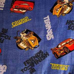 May include: Blue fabric with a repeating pattern of cartoon cars, including red and yellow vehicles. The fabric has the text "Friends to the Finish" and "Team 95" repeated throughout.
