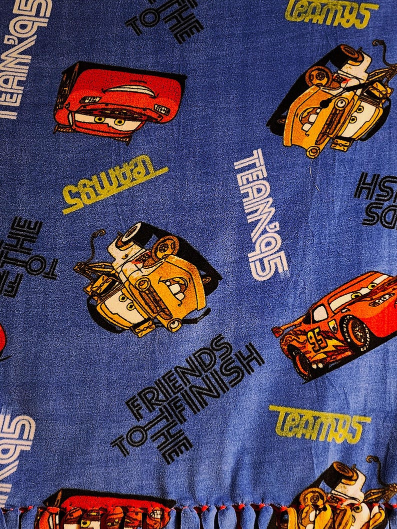 May include: Blue fabric with a repeating pattern of cartoon cars, including red and yellow vehicles. The fabric has the text "Friends to the Finish" and "Team 95" repeated throughout.