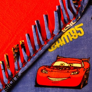 May include: A red fleece blanket with a blue Cars themed fabric on the other side. The Cars fabric features a red race car with a smiling face and the text "Cars 95". The blanket has a blue fringe around the edges.