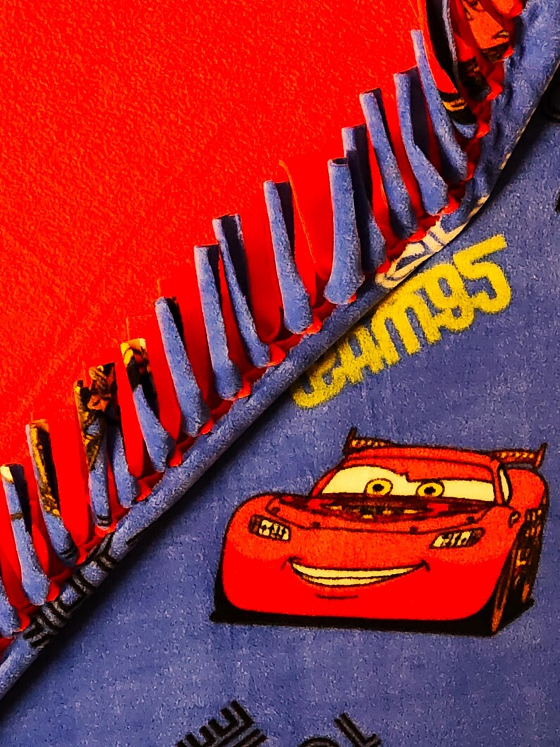 May include: A red fleece blanket with a blue Cars themed fabric on the other side. The Cars fabric features a red race car with a smiling face and the text "Cars 95". The blanket has a blue fringe around the edges.