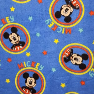 K&ouml;nnte beinhalten: Blauer Stoff mit einem sich wiederholenden Muster von Mickey Mouse in einem gelben, roten und orangen Kreis. Der Kreis hat das Wort "Mickey" darum herum geschrieben. &Uuml;ber den Stoff sind kleine gelbe und rote Sterne verteilt.