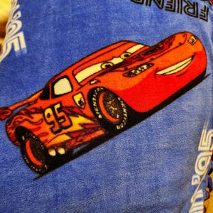 May include: A red and yellow cartoon race car with the number 95 on the side. The car is on a blue fleece blanket with the words "Friends" and "Lightning McQueen" printed on it.