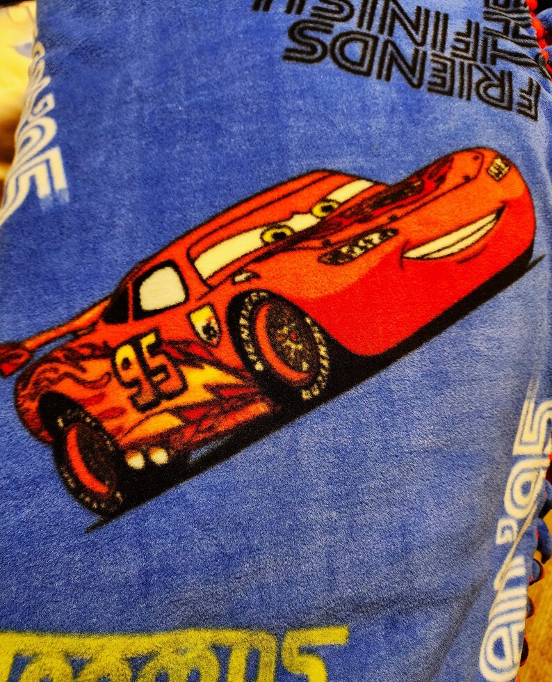 May include: A red and yellow cartoon race car with the number 95 on the side. The car is on a blue fleece blanket with the words "Friends" and "Lightning McQueen" printed on it.