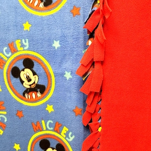 K&ouml;nnte beinhalten: Eine blaue Fleece-Decke mit rotem Fleece-Rand. Die Decke zeigt ein sich wiederholendes Muster von Mickey Mouse in einem gelben Kreis mit dem Wort "Mickey" auf der Decke. Die Decke hat einen roten Fransen am Rand.