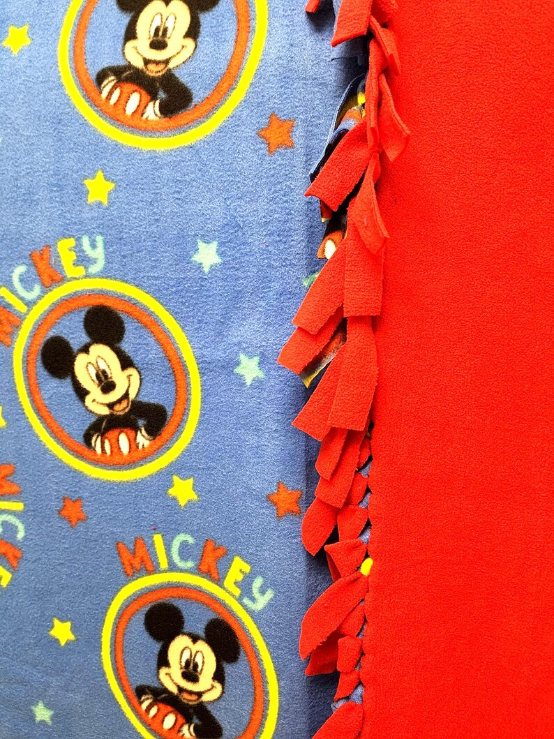 K&ouml;nnte beinhalten: Eine blaue Fleece-Decke mit rotem Fleece-Rand. Die Decke zeigt ein sich wiederholendes Muster von Mickey Mouse in einem gelben Kreis mit dem Wort "Mickey" auf der Decke. Die Decke hat einen roten Fransen am Rand.