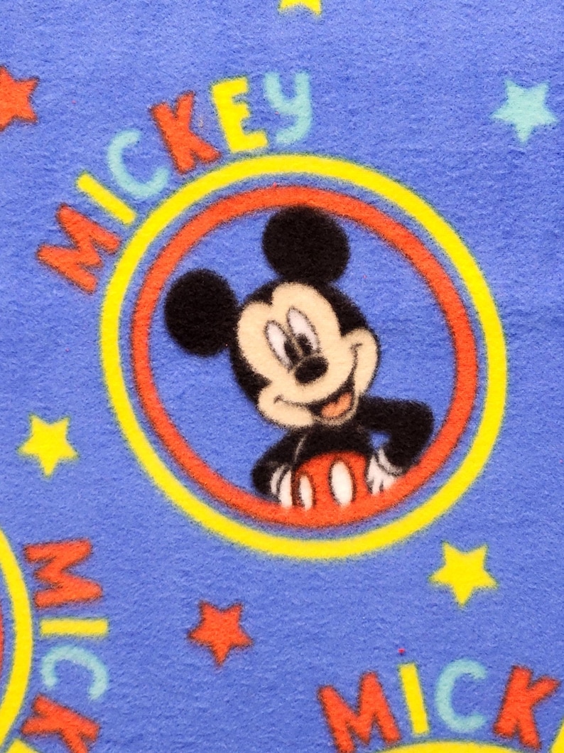 K&ouml;nnte beinhalten: Blauer Stoff mit einem sich wiederholenden Muster von Mickey Mouse in einem gelben und roten Kreis mit dem Wort "Mickey" in gelben Buchstaben. Der Hintergrund ist blau mit gelben Sternen.