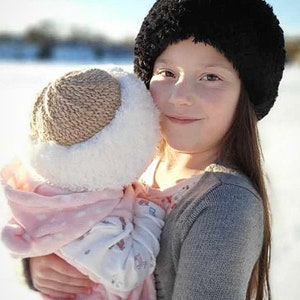 Könnte beinhalten: Eine junge Person mit einem schwarzen Pelzmützchen hält eine Puppe, die einen rosa-weißen Strickhut und einen rosa-weißen gemusterten Mantel trägt.