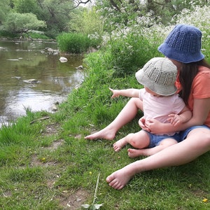 Peut inclure: Deux personnes, l'une portant un chapeau crocheté bleu et l'autre un chapeau crocheté beige, sont assises au bord d'une rivière. Elles regardent toutes les deux quelque chose au loin. La berge de la rivière est recouverte d'herbe verte.