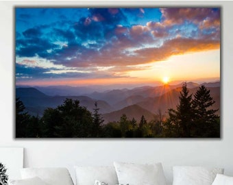 May include: A panoramic view of a mountain range at sunset. The sky is a vibrant mix of blue, pink, and orange with fluffy clouds. The sun is setting behind the mountains, casting long shadows and creating a warm glow. The mountains are covered in trees and are silhouetted against the sky.