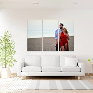 Puede incluir: Una impresi&oacute;n en lienzo dividida que presenta a una pareja con ropa roja y blanca de pie en una playa. La pareja mira hacia un lado, y el hombre tiene el brazo alrededor de la mujer. La playa es arenosa y el cielo es azul.