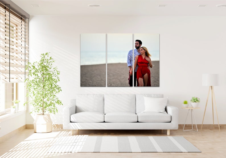 Puede incluir: Una impresi&oacute;n en lienzo dividida que presenta a una pareja con ropa roja y blanca de pie en una playa. La pareja mira hacia un lado, y el hombre tiene el brazo alrededor de la mujer. La playa es arenosa y el cielo es azul.