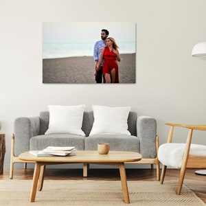 Puede incluir: Una pareja posa para una foto en una playa. El hombre lleva una camisa azul y la mujer lleva un vestido rojo. La playa es arenosa y el agua est&aacute; en el fondo.