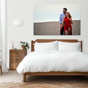 Puede incluir: Una pareja camina de la mano en una playa de arena. La mujer lleva un vestido rojo y el hombre lleva una camisa azul y blanca con estampado. La pareja est&aacute; enmarcada por una pared blanca y una cama blanca con ropa de cama blanca.