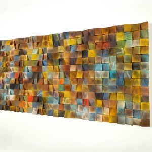 May include: A wall art piece made of wood with a mosaic of different colored wood squares. The squares are arranged in a wave pattern and are all different sizes. The colors are mostly shades of brown, yellow, and blue.