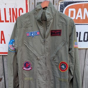 Könnte beinhalten: Ein olivgrüner Fluganzug mit Frontreißverschluss, langen Ärmeln und mehreren gestickten Aufnähern. Die Aufnäher umfassen ein Top Gun-Logo, ein Goose-Namensschild, eine Flagge der Vereinigten Staaten und verschiedene kreisförmige Luftfahrt-Embleme.