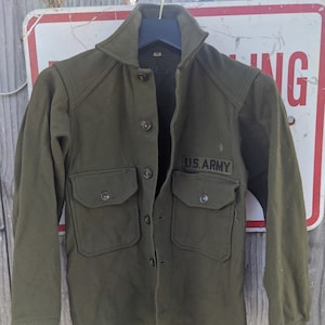 Puede incluir: Una chaqueta de camisa de lana verde con cierre de botones y dos bolsillos de solapa. La chaqueta tiene un parche negro "U.S. ARMY" en el pecho izquierdo.