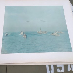 May include: A black and white photograph of a fleet of ships at sea. The ships are sailing in formation, with a large aircraft carrier in the center. Several airplanes are flying overhead.