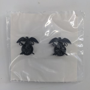 May include: Two black metal eagle and cannon insignia badges in a clear plastic bag on a white background.