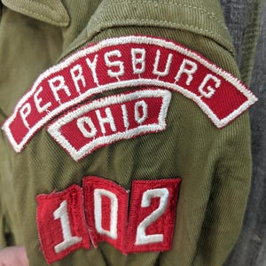 May include: Close-up of an olive green jacket sleeve with three embroidered patches. The top patch reads "PERRYSBURG OHIO" in white letters on a red background. Below are two patches with the number "102" in white on a red background.