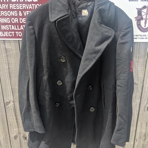 Op de afbeelding: Een zwarte wollen pea coat met een dubbele rij knopen, vier zichtbare knopen en een revers. De jas hangt aan een houten kleerhanger tegen een verweerde houten schutting. De jas heeft een label aan de binnenkant.