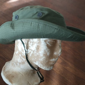 May include: Olive green boonie hat with a wide brim and two black snap closures. The hat has a chin strap and is displayed on a mannequin head. The hat is made of a durable fabric.