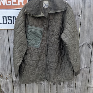 May include: Olive green quilted jacket with a corduroy collar and a single chest pocket. The jacket has a full-length zipper and ribbed cuffs. The jacket is hanging on a wooden fence.