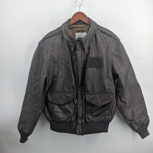 Puede incluir: Chaqueta bomber de cuero marrón con cuello y dos bolsillos delanteros. La chaqueta tiene dobladillo y puños acanalados. El nombre de la marca "SADDLELY" es visible en el cuello. La chaqueta está colgada en una percha de madera.