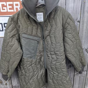 May include: Olive green quilted jacket with a high collar and a zip-up front. The jacket features a chest pocket and ribbed cuffs. A tag with text is visible on the upper chest. The jacket is hanging on a metal hanger.