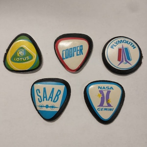 May include: A collection of five vintage automotive and space-themed pins. The pins feature logos for Lotus, Cooper, Plymouth, Saab, and NASA Gemini. Each pin has a unique shape and color scheme, with a black border.