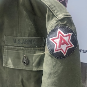 May include: Olive green military jacket with a pocket and the text "U.S. ARMY". The jacket features a patch with a red star and the letter "A" on the sleeve. The jacket is made of a durable fabric.