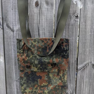 Könnte beinhalten: Eine Tragetasche mit Tarnmuster und olivgrünen Trägern. Die Tasche hat einen Klappenverschluss mit zwei Metallverschlüssen. Das Tarnmuster weist Grün-, Braun- und Schwarztöne auf. Die Tasche hängt an einem transparenten Kunststoffbügel.