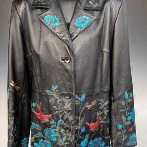 May include: Black leather jacket with floral and bird embroidery. The jacket features a button-up front, a notched collar, and detailed turquoise and red floral and bird designs. The jacket is a medium length.