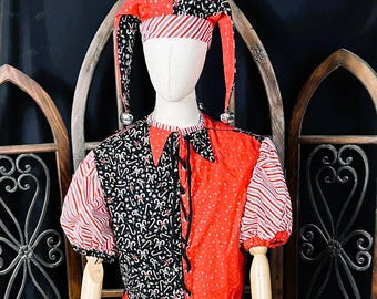 May include: A red and black jester costume displayed on a mannequin. The costume features a pointed hat with bells, a collared top, and a skirt. The top is red with white polka dots and black with a candy cane pattern. The sleeves are red and white striped.