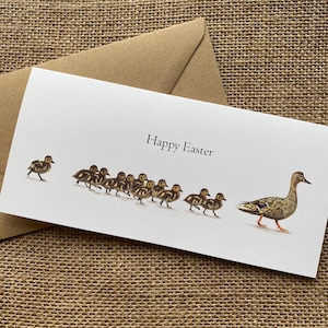 Peut inclure: Une carte de Pâques blanche avec l'inscription "Happy Easter" présente une illustration à l'aquarelle d'une cane et de ses canetons. Les canetons suivent la cane. Une enveloppe marron est derrière la carte.