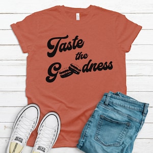 May include: A rust-coloured t-shirt with the text "Taste the Goodness" in black script with two black macarons below the text.