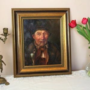 May include: A framed oil painting of a man with a beard and mustache, wearing a dark hat and a brown jacket. He is holding a pipe in his mouth and a glass in his hand. The painting is set against a dark background.