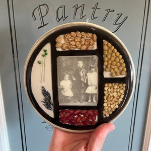 May include: A round, white ceramic plate with a black border and six compartments. Each compartment contains a different type of dried beans or seeds. A black and white photo of three children is in the center compartment. The plate has the word "Pantry" written on it.