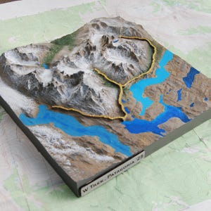 May include: A 3D topographical model of a mountain range with blue water features and a gold-colored trail. The model is set on a map and has the text "W Trek - Patagonia 25". The mountains are white and gray.