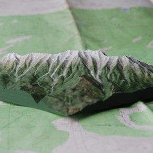 May include: A detailed, three-dimensional wooden mountain range model with green slopes and white, snow-capped peaks. The model sits on a topographic map, showcasing the terrain's features. The base of the model is painted green.