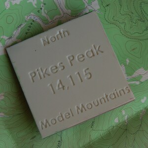 K&ouml;nnte beinhalten: Ein wei&szlig;es Schild mit dem Text "North Pikes Peak 14,115 Model Mountains" auf einer gr&uuml;nen topografischen Karte.