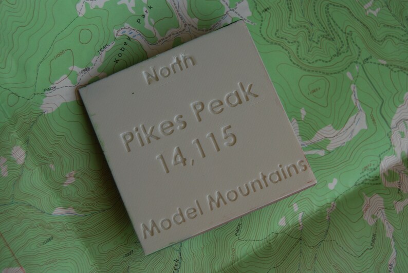 K&ouml;nnte beinhalten: Ein wei&szlig;es Schild mit dem Text "North Pikes Peak 14,115 Model Mountains" auf einer gr&uuml;nen topografischen Karte.