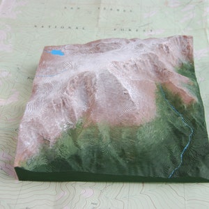 K&ouml;nnte beinhalten: Ein 3D-Modell einer Bergkette mit schneebedeckten Gipfeln und gr&uuml;nen T&auml;lern. Das Modell besteht aus einem strukturierten Material und ist in Braun-, Gr&uuml;n- und Wei&szlig;t&ouml;nen bemalt.